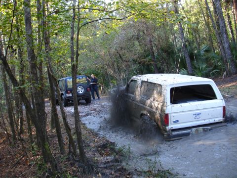 1989 Ford Bronco - A white Ford Bronco kicks up mud while driving through a wooded trail, with a Jeep parked nearby.