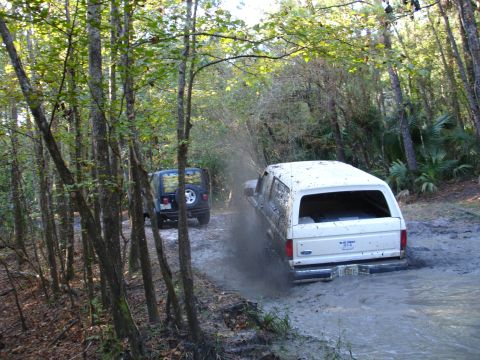 1989 Ford Bronco - A white 1989 Ford Bronco navigates a muddy trail, kicking up spray as a Jeep follows behind.