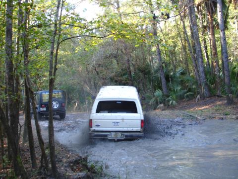 1989 Ford Bronco - A white SUV navigates a muddy path in a wooded area, with another vehicle visible ahead.