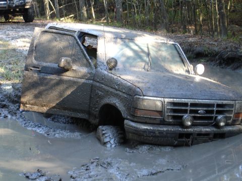 1989 Ford Bronco - Ford Bronco stuck deep in mud with driver's door ajar; another vehicle is in the background.