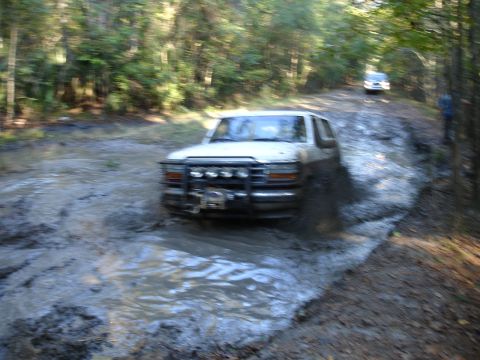 1989 Ford Bronco - A white Ford Bronco navigates a muddy path in a wooded area, with another vehicle visible in the distance.