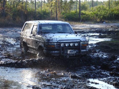 1989 Ford Bronco - White SUV navigating a muddy track, splashing through the mire.