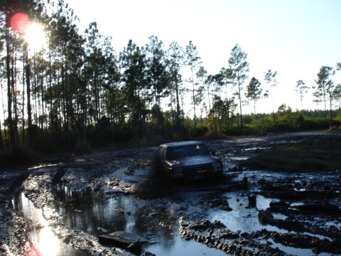 1989 Ford Bronco - A 1989 Ford Bronco navigates a muddy off-road course, with trees lining the horizon.