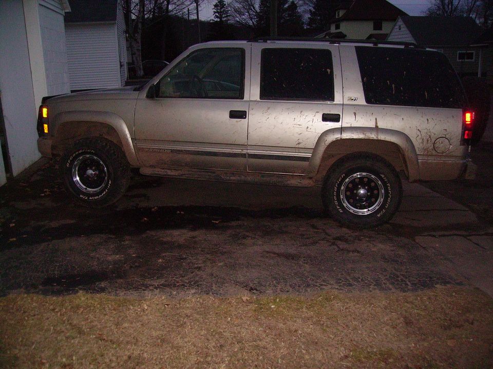 Gold Chevrolet Tahoe parked on a dirt driveway, side view. Mud splatters on body