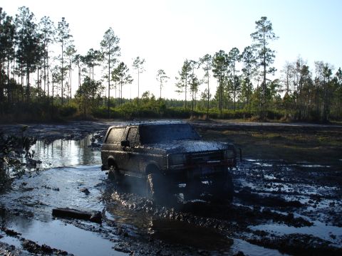 1989 Ford Bronco - A 1989 Ford Bronco navigates a challenging muddy terrain, with trees in the background.