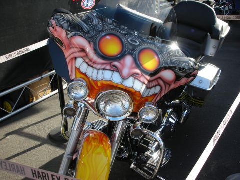 1998 Ford Crown Victoria - Front of a Harley-Davidson motorcycle with an airbrushed grinning demon face and goggles painted on the fairing under NHRA signage.
