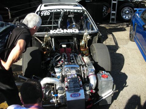 1998 Ford Crown Victoria - man inspects the exposed twin-turbo engine bay of a black Mazdaspeed drag car with Konig and NHRA decals in the pits.