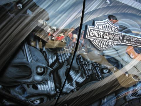 1998 Ford Crown Victoria - Airbrushed skull and weapon artwork on a vehicle body beside chrome Harley-Davidson and F-150 Supercharged emblems.