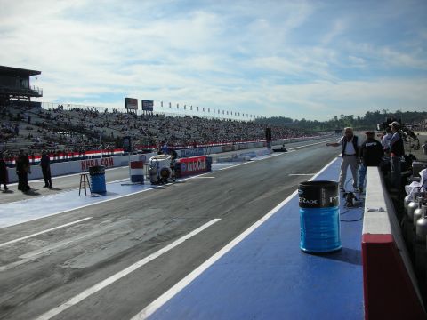 1998 Ford Crown Victoria - drag strip starting line with painted blue surface, grandstands, and NHRA.com signage under a partly cloudy sky.