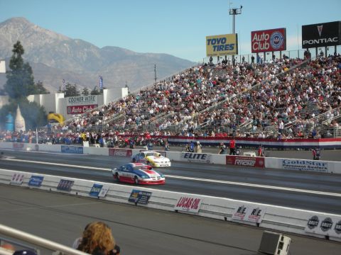 1998 Ford Crown Victoria - Two Pro Stock drag cars launch side by side down a strip lined with sponsor banners, full grandstands, and mountains beyond.