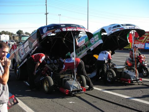 1998 Ford Crown Victoria - Two nitro Funny Cars sit in the pits with bodies raised as crews service the engines, wearing Auto Club and GTX livery.