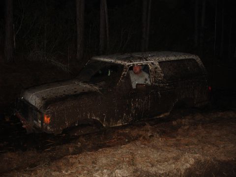1989 Ford Bronco - A mud-covered SUV is stuck in a wooded area at night, with a driver visible inside.