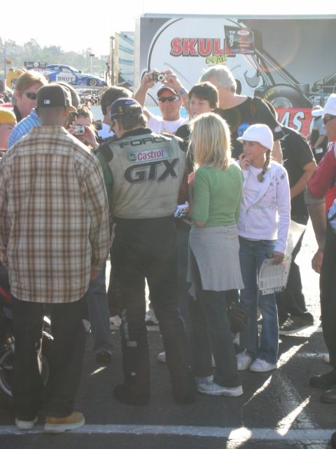1998 Ford Crown Victoria - Spectators take photos of a driver wearing a Ford Castrol GTX firesuit near a Skull Gear vendor display.