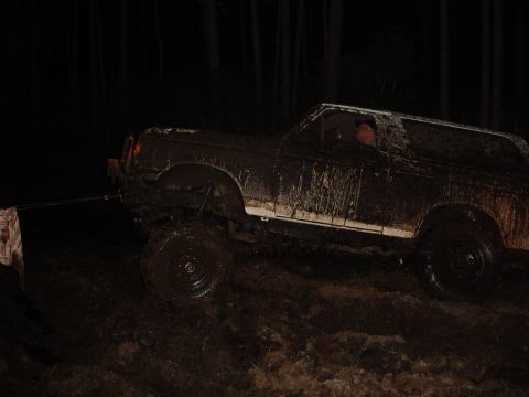 1989 Ford Bronco - A muddy SUV is stuck in a dark, wooded area, with a tow strap attached to the front.