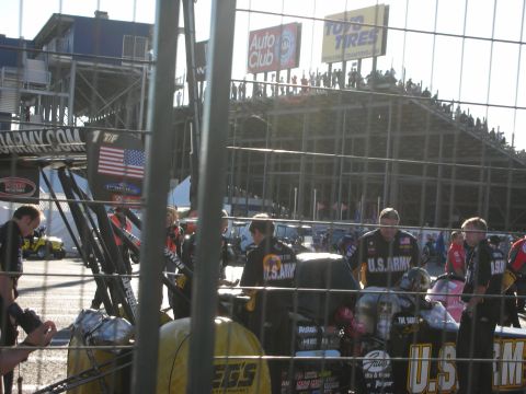 1998 Ford Crown Victoria - Crew members around a U.S. Army-sponsored Top Fuel team setup in the pits, with grandstands and trackside signage behind.