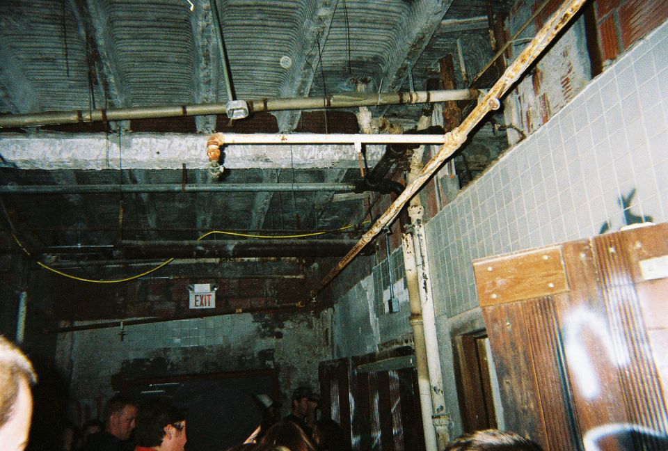 Dark grungy hallway with exposed pipes, tiled wall, EXIT sign, and a crowd of people