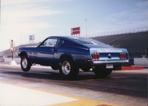 1965 Ford Mustang - A blue Ford Mustang lifts its front wheels during a drag race, showing off its rear end and racing slicks.