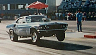 1965 Ford Mustang - A modified 1969 Ford Mustang drag car competes on a track, kicking up dust as its front wheels leave the pavement.