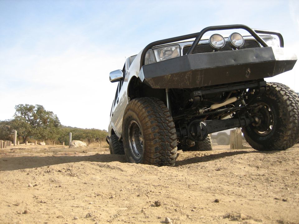 Silver SUV with large off-road tires and a black bull bar, angled from the front-right, driving on a dirt track