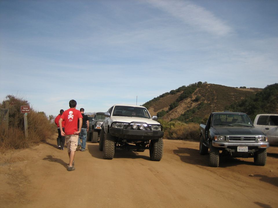 Several off-road trucks and SUVs parked on a dirt road with hills in the background. One truck has a "SUPERMOTORS" logo