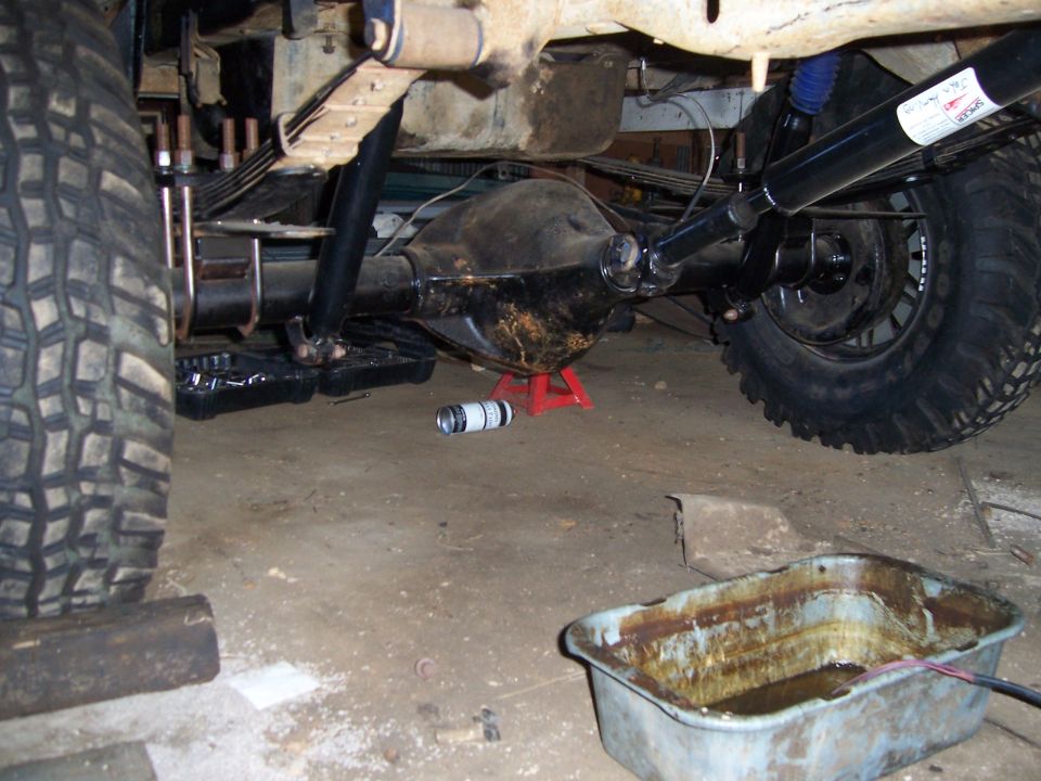 Undercarriage view of a vehicle's rear axle, differential, leaf springs, and shock absorber, with a drain pan and tools nearby
