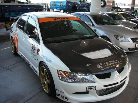 1998 Ford Crown Victoria - white Mitsubishi Lancer Evolution with a carbon-fiber hood and sponsor decals parked indoors beside other cars.