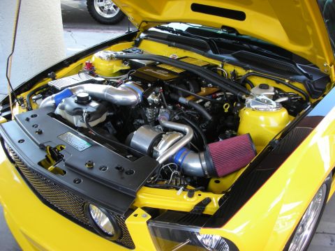 1998 Ford Crown Victoria - Engine bay of a yellow car fitted with a supercharger, polished piping, and a red cone intake filter under an open hood.
