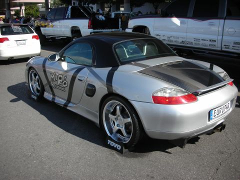1998 Ford Crown Victoria - silver Porsche Boxster with a two-tone graphic wrap and rear spoiler parked on a Toyo tire mat at a show.