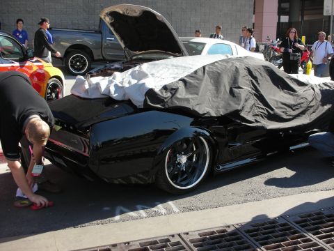 1998 Ford Crown Victoria - man crouches to detail a black lowered car partly draped in a car cover at a busy outdoor event.