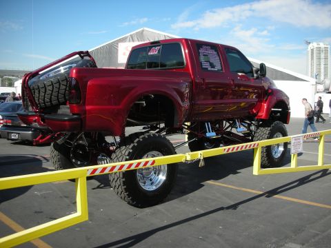 1998 Ford Crown Victoria - deep red lifted crew-cab truck with flame graphics raised on a tall suspension, shown behind yellow barriers.