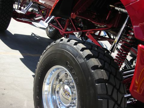 1998 Ford Crown Victoria - polished alloy wheel with chunky off-road tire sits under a red lifted truck with red tubular framework and coilover shocks.