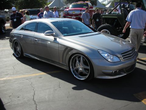 1998 Ford Crown Victoria - lowered silver Mercedes-Benz CLS four-door coupe on chrome split-spoke wheels parked among a crowd at a car show.