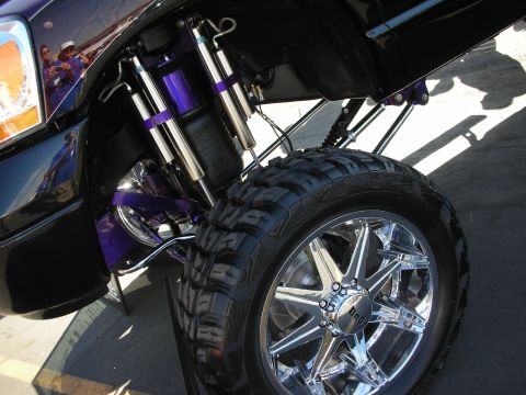 1998 Ford Crown Victoria - Front suspension of a lifted truck with purple painted arms, twin reservoir shocks, and a chrome wheel with mud tire.