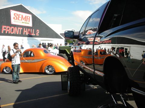 1998 Ford Crown Victoria - orange chopped hot rod coupe parked next to a black lifted truck outside a tent marked SEMA Show Front Hall 2.