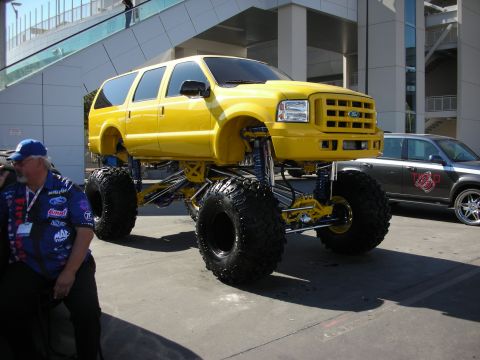 1998 Ford Crown Victoria - yellow lifted Ford Excursion SUV towers on massive off-road tires and exposed suspension in an outdoor show lot.