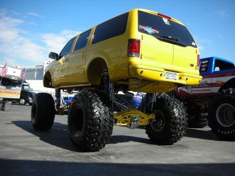 1998 Ford Crown Victoria - Rear three-quarter of a yellow lifted Ford Excursion on huge tires next to a red Firestone-liveried monster truck.