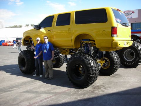 1998 Ford Crown Victoria - Two men pose next to a towering yellow lifted Ford Excursion SUV on enormous off-road tires in an outdoor show lot.