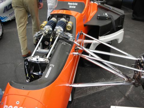1998 Ford Crown Victoria - Close-up of an orange open-wheel race car's front pushrod suspension with chrome rockers, coilovers, and a Do Not Sit notice.