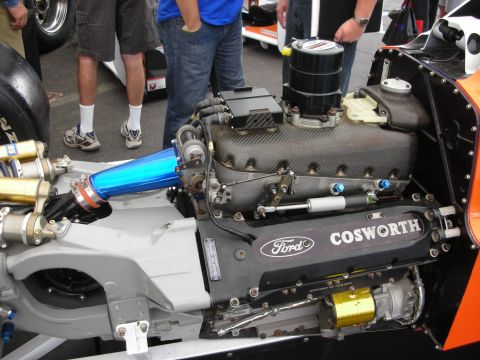 1998 Ford Crown Victoria - Ford Cosworth V8 race engine sits exposed in an open-wheel car as spectators stand around it at a track.