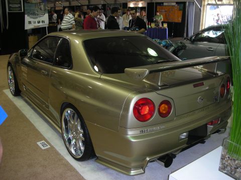 1998 Ford Crown Victoria - gold Nissan Skyline coupe with a rear wing and chrome wheels is displayed at an indoor car show booth.