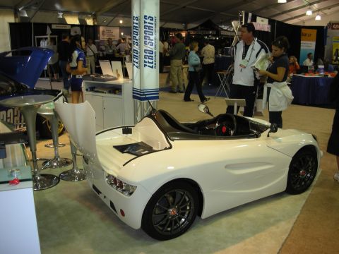 1998 Ford Crown Victoria - small white open-cockpit roadster concept car sits on display at a Zero Sports booth in a busy exhibition hall.