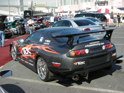 1998 Ford Crown Victoria - black Toyota Supra with orange flame graphics and a tall rear wing sits in an outdoor automotive show lot.