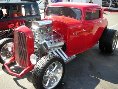 1998 Ford Crown Victoria - red three-window hot rod coupe sits with an exposed chromed V8, headers, and polished wheels at an outdoor show.