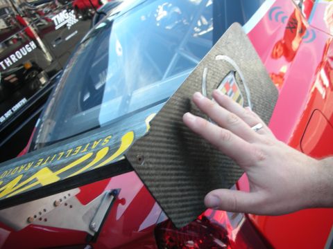 1998 Ford Crown Victoria - hand holds a carbon-fiber plate bearing a Cadillac crest beside a red race car at an outdoor event.