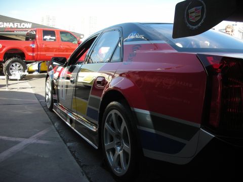 1998 Ford Crown Victoria - rear side of a graphically wrapped Cadillac with a tall rear wing is parked beside a red truck at a show.