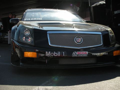 1998 Ford Crown Victoria - black Cadillac CTS race car shows its mesh grille, Cadillac crest, and Mobil 1 and SCCA Pro Racing decals in a garage.