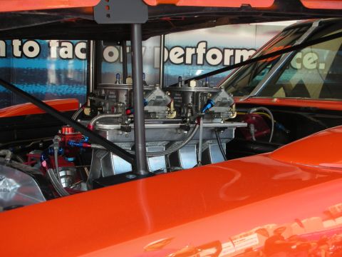 1998 Ford Crown Victoria - twin-carburetor tunnel-ram intake with braided lines sits in the engine bay of an orange race car, viewed through the windshield.
