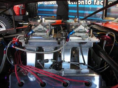 1998 Ford Crown Victoria - Close-up of two four-barrel carburetors atop a polished intake manifold, fed by braided fuel lines with red plug wires below.