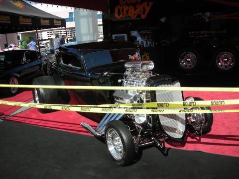 1998 Ford Crown Victoria - black hot rod with an exposed supercharged V8 and zoomie headers sits on red carpet behind yellow caution tape at a show.