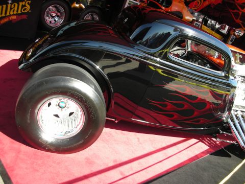 1998 Ford Crown Victoria - black flamed hot rod coupe sits on a red carpet display with a polished alloy wheel and wide rear racing slick.